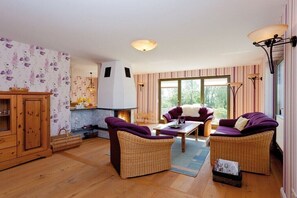 Un salon chaleureux, avec des meubles en osier, une cheminée et une vue dégagée grâce à de grandes fenêtres.