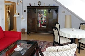 Un salon avec un canapé rouge, une table basse en verre, des fauteuils à motifs, un meuble en bois avec des portes en verre et une table blanche avec une lampe.