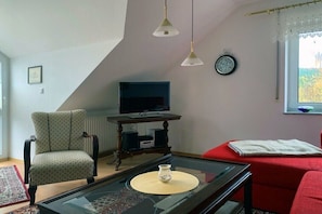 A living room with a television, a red sofa, a patterned armchair, a glass coffee table, and a clock on the wall. - Holiday apartment with 1 bedroom (Seebad Ahlbeck)