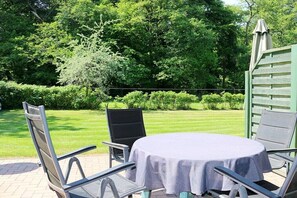 Een terras buiten met een ronde tafel en stoelen, omringd door groen en een hek.