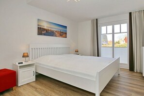 Un dormitorio con una cama grande, una mesita de noche, un otoman rojo, una ventana con cortinas y un cuadro en la pared.