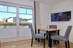 Ein Zimmer mit einem Esstisch, zwei Stühlen, einem an der Wand befestigten Fernseher und einem großen Fenster mit Jalousien.