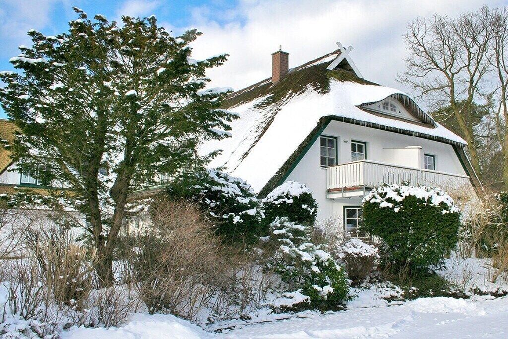 A snow-covered house with a steep roof, surrounded by snow-laden trees and bushes.