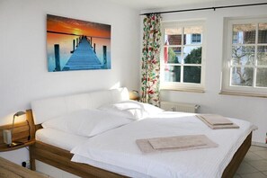 Een slaapkamer met een houten bed, wit beddengoed, een nachtkastje met een lamp, een raam met gordijnen en een schilderij van een zonsondergang boven een pier.