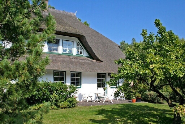 Een traditioneel huis met rieten dak, een witte gevel, meerdere ramen en een verzorgde tuin met bomen en struiken.