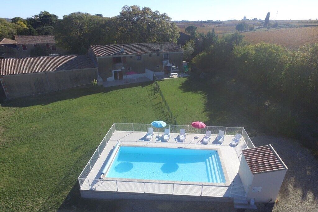 Piscina com espreguiçadeiras e guarda-sóis, cercada por área gramada e um edifício ao fundo.