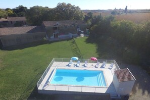 Una piscina con sillones de descanso y sombrillas, rodeada por un área cercada con césped y un edificio al fondo.