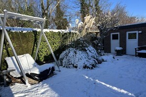 A snow-covered outdoor area with a swing set, a bush, and a brick building with a door. - Holiday home with 3 bedrooms (BX Herkingen)