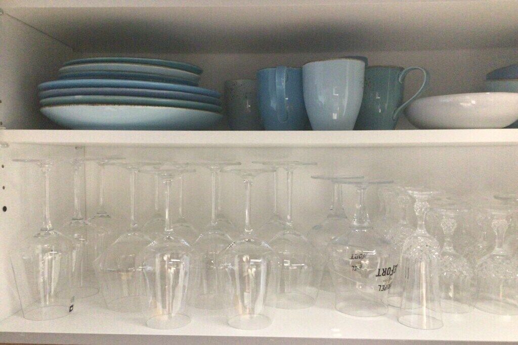 A shelf with stacked plates and bowls, and a row of wine glasses below.