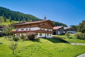 Un chalet traditionnel doté d’un balcon orné de fleurs, niché dans une vallée verdoyante et luxuriante, avec une chaîne de montagnes en toile de fond.
