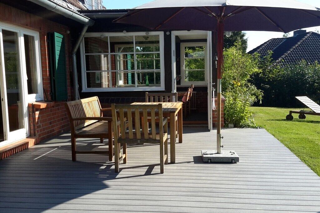 En træterrasse med et bord og stole, en parasol og et murstenshus med store vinduer.