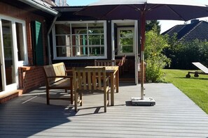 A wooden deck with a table and chairs, a patio umbrella, and a brick house with large windows. - Holiday home with 2 bedrooms (Sierksdorf)