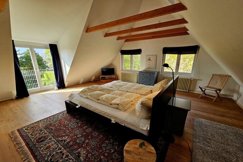 Ein Schlafzimmer mit schrägverdeckter Decke, einem Bett, einer Kommode, einem Sessel und einem Fenster mit Aussicht.