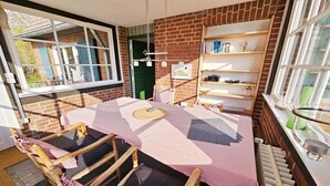 Ein gemütliches Zimmer mit Backsteinwand, einem Fenster, einem Tisch mit kariertem Tischlaken und einem Bücherregal.