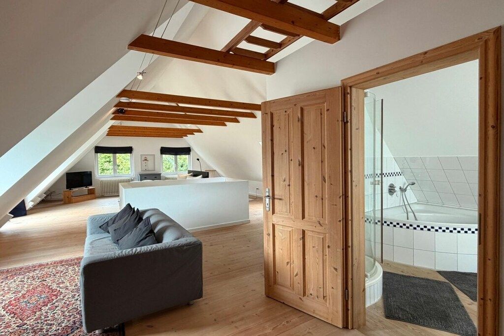 A modern interior with a loft-style ceiling, a grey sofa, and a bathroom with a glass shower enclosure.
