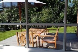 An outdoor patio with a wooden table and chairs, a white umbrella, and a view of a green lawn and trees. - Holiday home with 2 bedrooms (Sierksdorf)