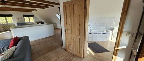 A room with a wooden ceiling, a grey sofa, a wooden door, and a view of a tiled area.