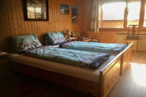 Een slaapkamer in een houten blokhut met twee bedden, een stoel, een tafel en een raam met gordijnen.