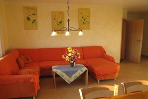 A living room with a sectional sofa, a small table with a vase of flowers, and a chandelier. - Bioland farm Teufelsberg (Horgenzell)
