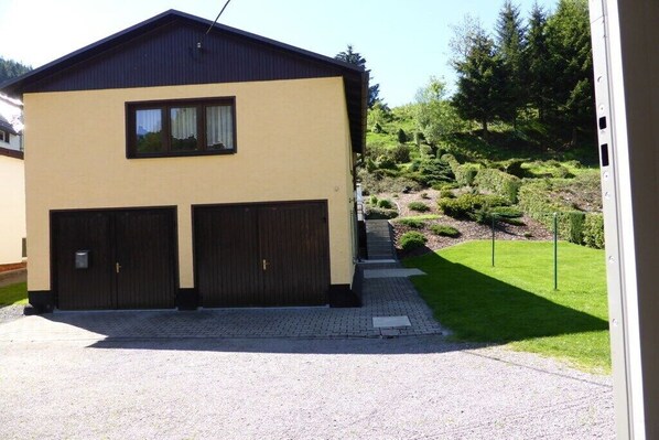 Een tweelaags gebouw met een garage, een verharde oprit en een verzorgd gazon met vorm gesnoeide struiken en bomen.