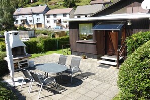 Een terras buiten met een tafel, stoelen, een barbecue en een houten gebouw met een raam.