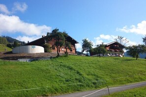 Paisatge rural amb una gran estructura cilíndrica, un edifici de fusta amb teulat inclinat i un edifici més petit amb teulat vermell.