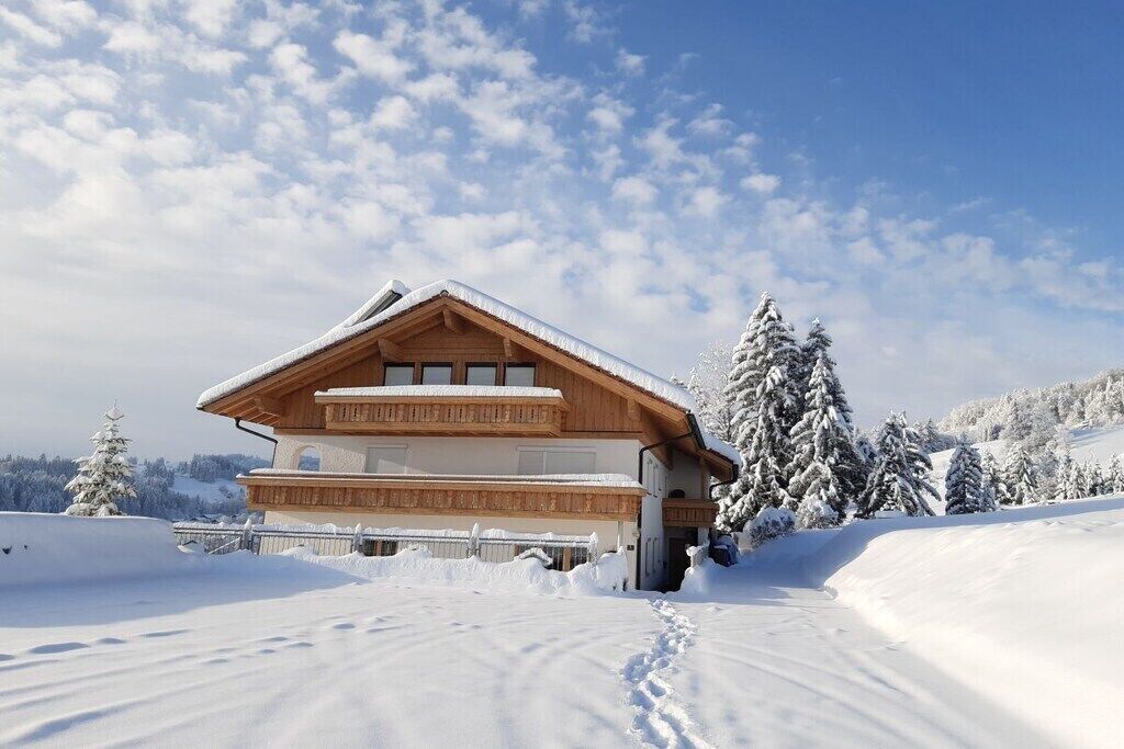A wooden chalet with a sloped roof, surrounded by snow-covered trees and a clear sky.