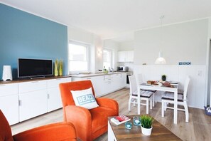 Ein modernes Wohnzimmer mit Fernseher, orangefarbenem Sessel und einem Essbereich mit Tisch und Stühlen.