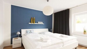 A modern bedroom with a large bed, bedside tables, a hanging light fixture, and a window with curtains. - TRINCHEN in the home by the sea (Büsum)