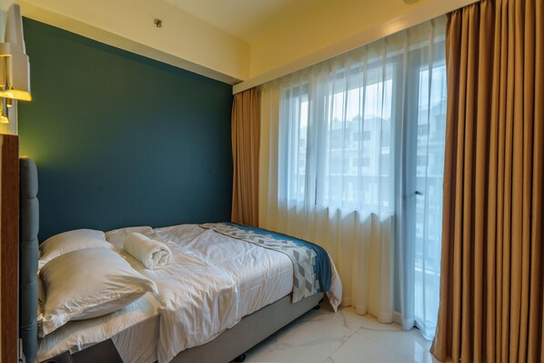 A hotel room with a bed, a headboard, a window with curtains, and a wall with a light switch.