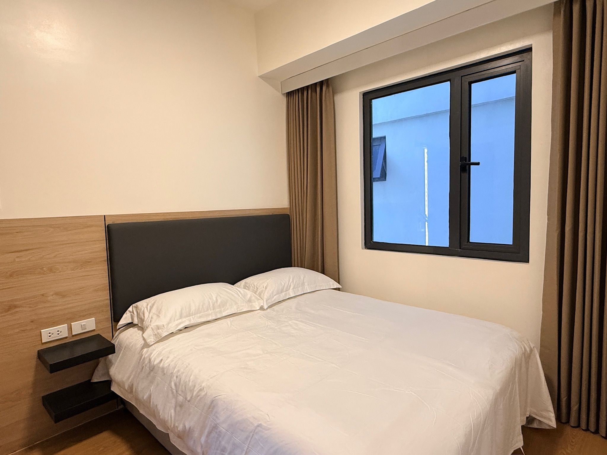 Una habitación de hotel con cama, mesita de noche, ventana con cortinas y vistas al exterior.