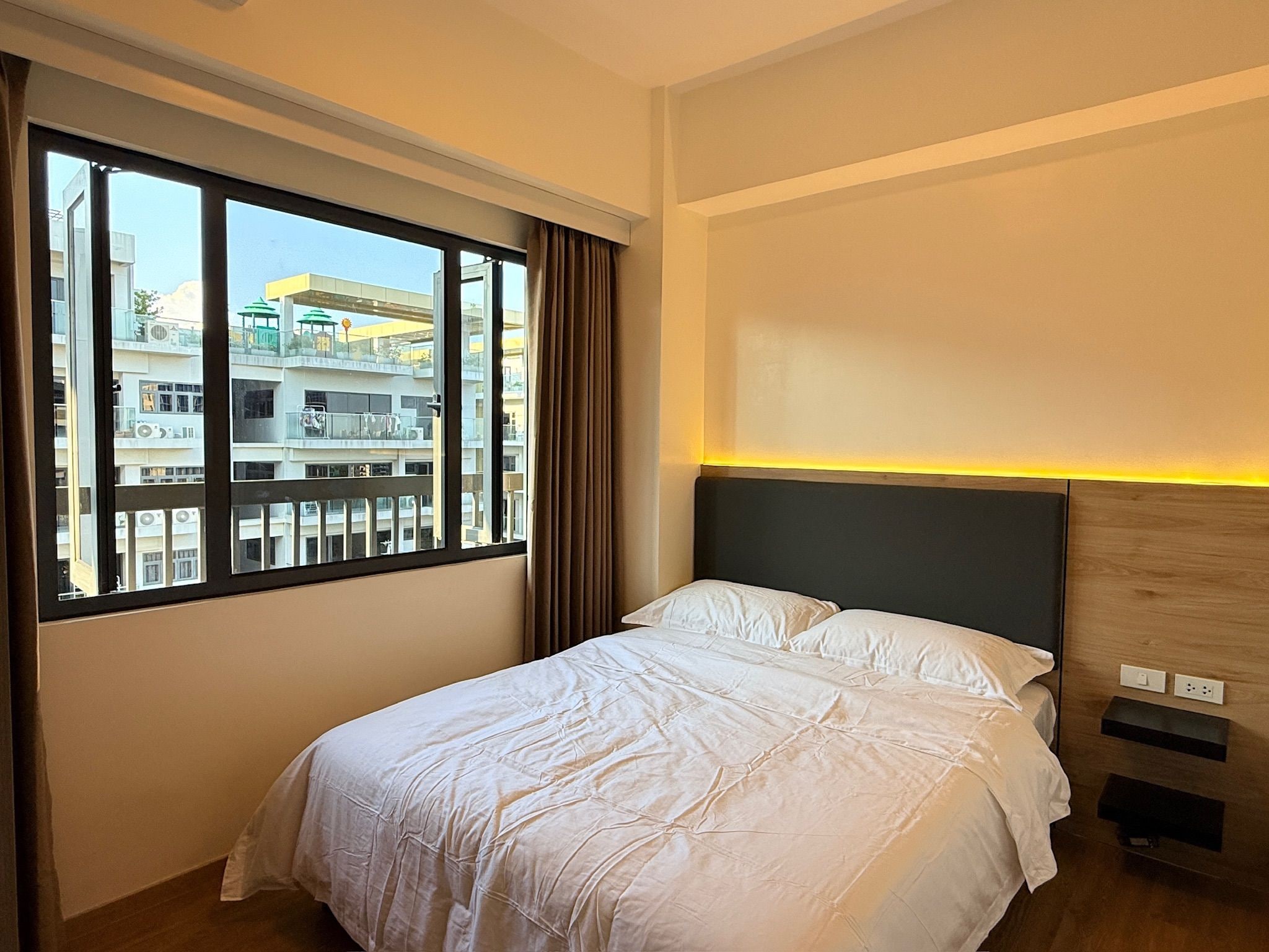 Habitación de hotel con cama, ventana que ofrece vistas a edificios, y una lámpara fijada a la pared.