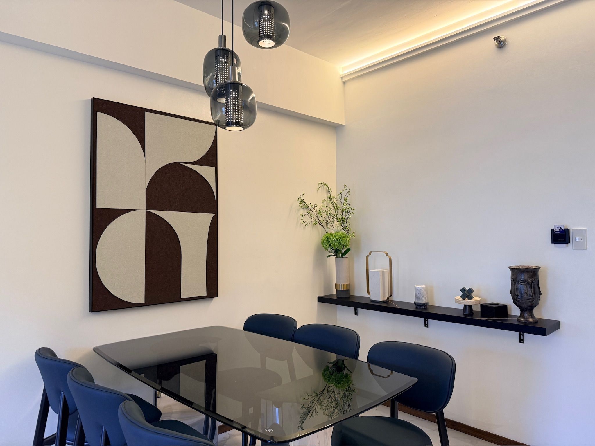 Uma área de refeições moderna com mesa de vidro, cadeiras azuis, pintura abstrata geométrica e um aparador com objetos decorativos.