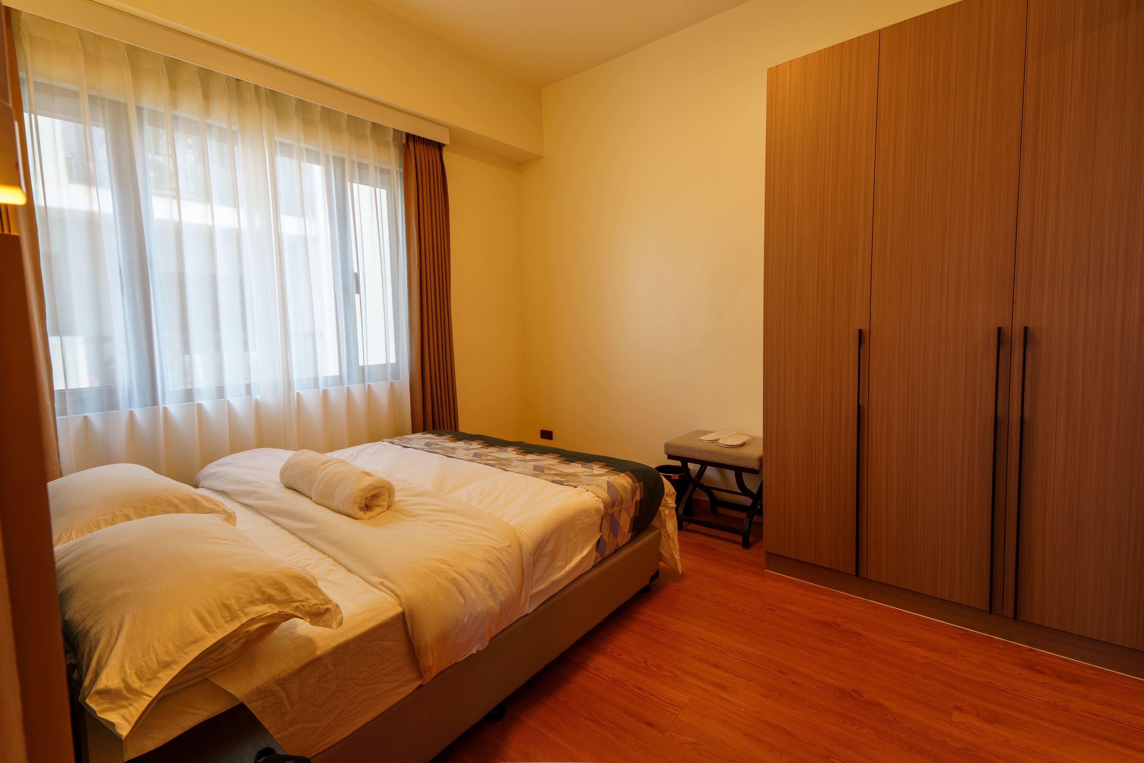 Quarto com cama, guarda-roupa de madeira, uma mesinha e janela com cortinas.