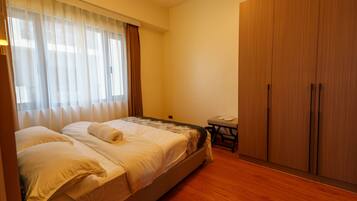 Quarto com cama, guarda-roupa de madeira, uma mesinha e janela com cortinas.