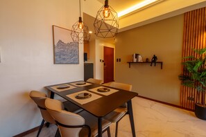 A dining area with a table set for four, chairs, pendant lights, a wall-mounted shelf with books, and a potted plant.