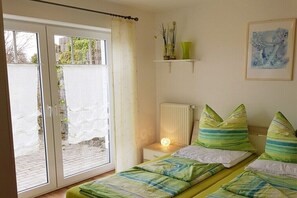 Un dormitorio con cama, mesita de noche con lámpara, radiador, ventana con cortinas y estante en la pared con objetos decorativos.