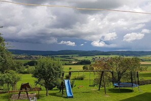 Un terrain de jeux équipé d’un toboggan, de balançoires et d’un trampoline, le tout situé dans un cadre verdoyant avec des champs et des collines à l’horizon.