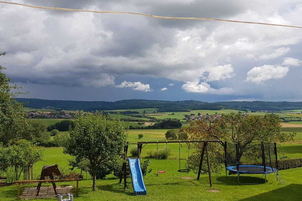 A playground with a slide, swings, and a trampoline, set against a backdrop of green fields and distant hills.