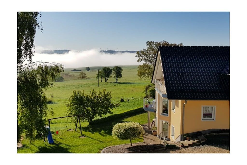 Una casa con balcón que da a un campo verde y un paisaje brumoso.