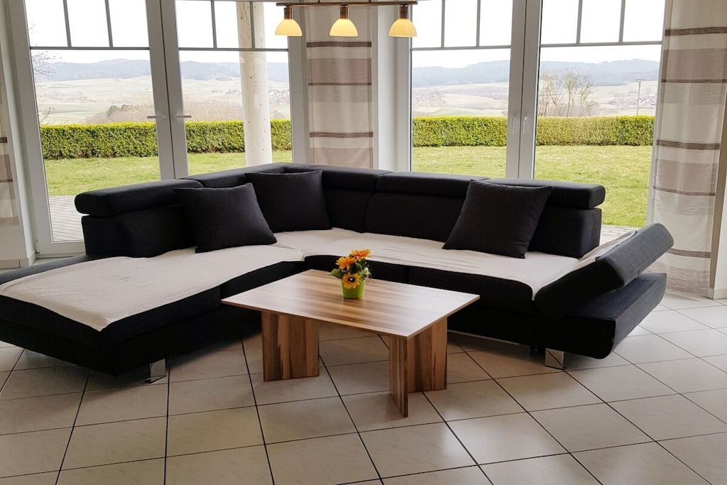 Ein modernes Wohnzimmer mit einem Ecksofa, einem Holztisch und Blick auf eine grüne Landschaft durch große Fenster.