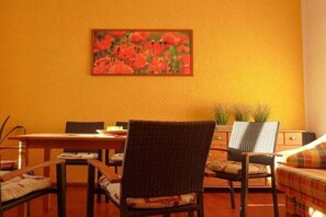 A dining area with a table and chairs, a painting of flowers on the wall, and a bed with a colorful blanket. - A1 in the apartment building near Müritz (Waren (Müritz))