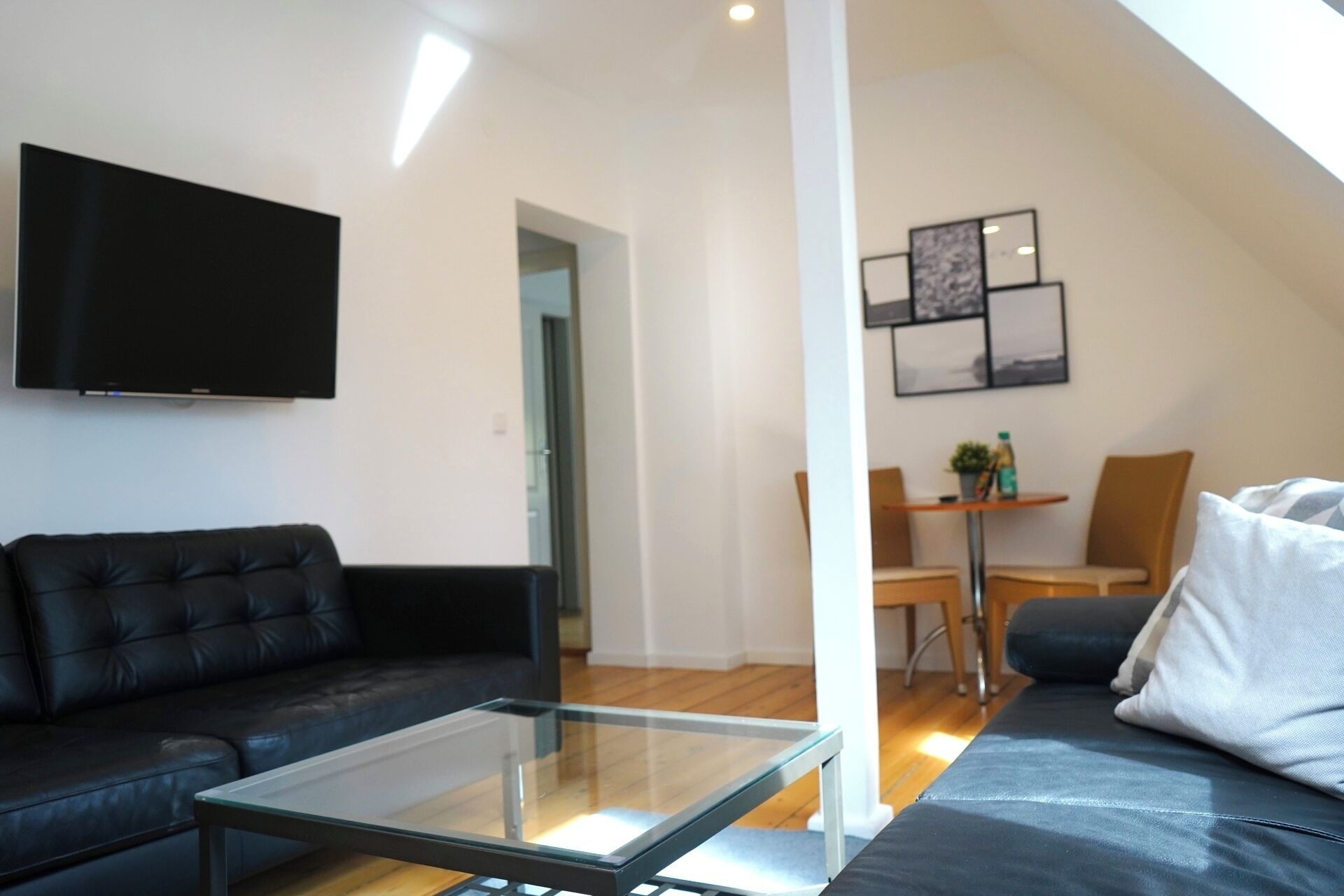 A modern living room with a glass coffee table, a black leather sofa, a flat-screen TV mounted on the wall, and a dining area with a small table and chairs.