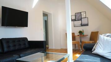 A modern living room with a glass coffee table, a black leather sofa, a flat-screen TV mounted on the wall, and a dining area with a small table and chairs.