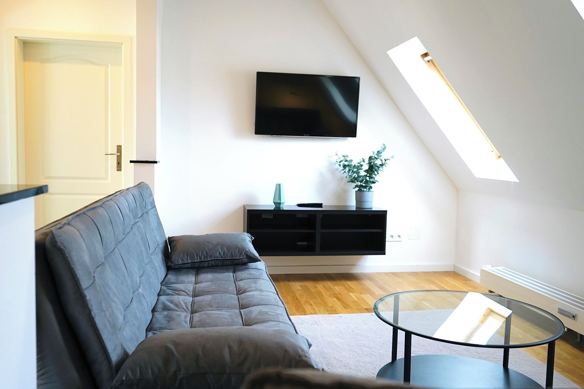 A modern living room with a sofa, a glass coffee table, a flat-screen TV mounted on the wall, and a skylight.