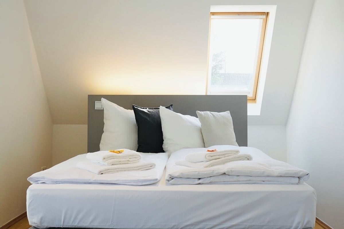 A neatly made bed with white linens and pillows, a dark headboard, and a small window with a view.