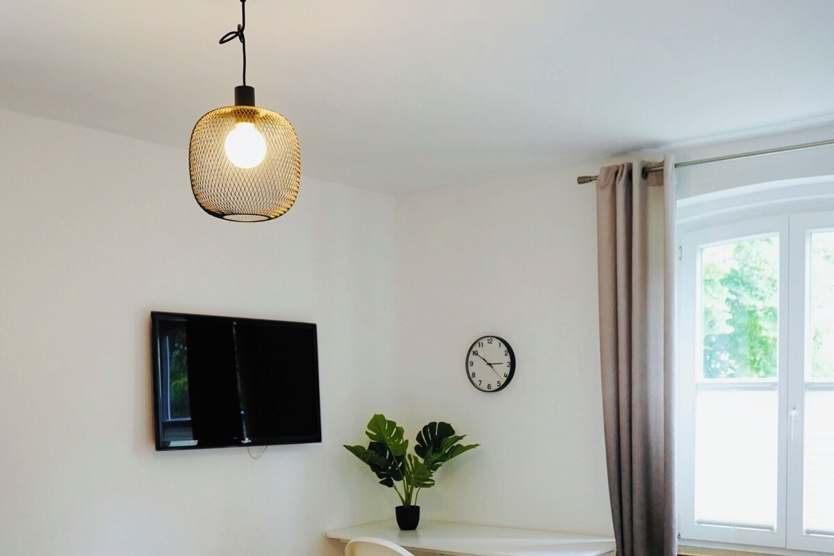 A bedroom with a bed, a desk, a television, a clock, and a window with curtains.