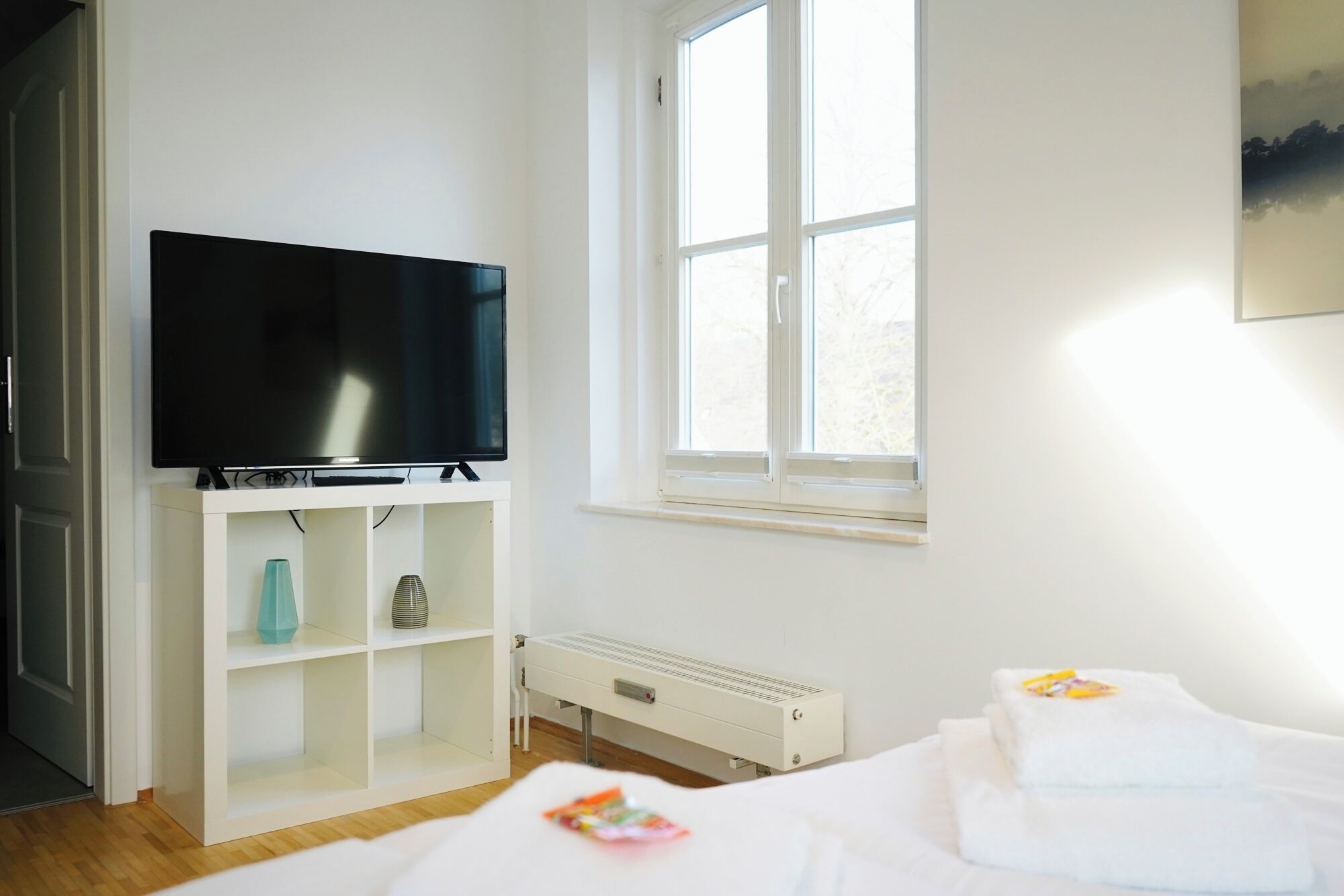 A hotel room with a flat-screen TV on a white stand, a window with a view of trees, a white radiator, and a bed with a white duvet.
