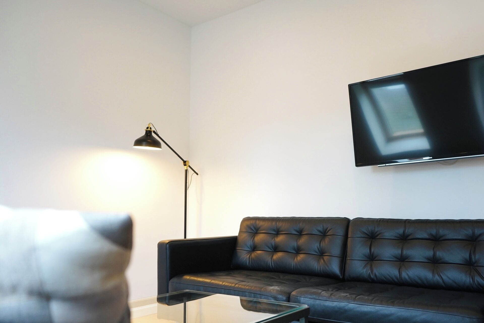 Un salon avec un canapé en cuir noir, une table basse en verre, un lampadaire noir et un téléviseur à écran plat fixé au mur.
