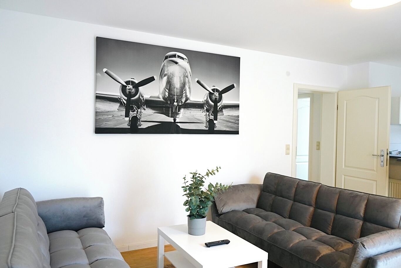 Ein Wohnzimmer mit einem großen schwarz-weiß Gemälde eines Flugzeugs, einem grauen Sofa, einem weißen Couchtisch mit Pflanze und Fernbedienung sowie einer Tür im Hintergrund.